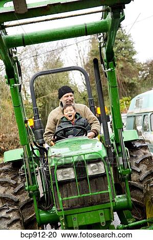 Stock Image - Boy driving a tractor with assistance of male adult. Fotosearch