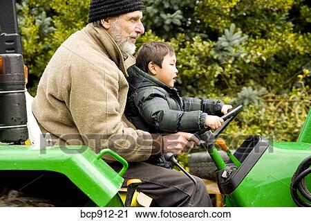 Stock Image - Boy driving a tractor with assistance of male adult. Fotosearch