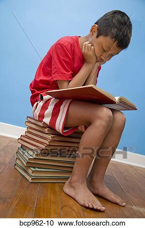 Stock Image - Boy reading while seated on stack of books. Fotosearch