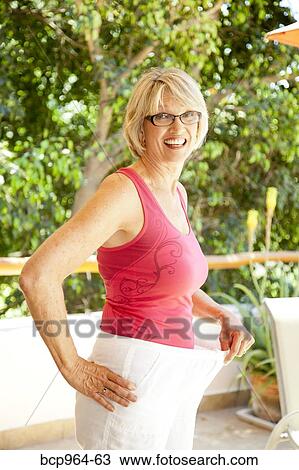 Stock Image - Happy woman holding out waistband of too large pants. Fotosearch
