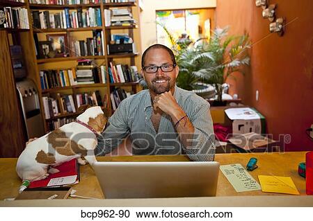 Stock Image - Man seated at home office with dog on desk. Fotosearch