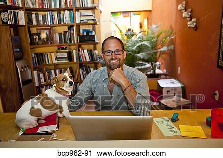 Stock Image - Man seated at home office with dog on desk. Fotosearch