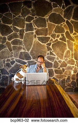 Stock Image - Man seated at long table with laptop and dog. Fotosearch