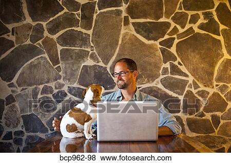 Stock Photo - Man seated at long table with laptop and dog. Fotosearch