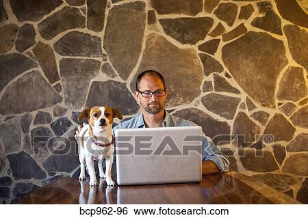 Man seated at long table with laptop and dog View Large Photo Image Stock Photograph - Man seated at long table with laptop and dog. Fotosearch