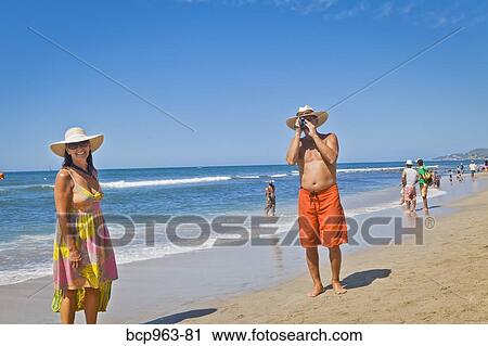 Stock Image - Man taking photograph of smiling woman on beach. Fotosearch