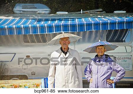 Stock Photography - Senior couple in rain hats near airstream camper. Fotosearch