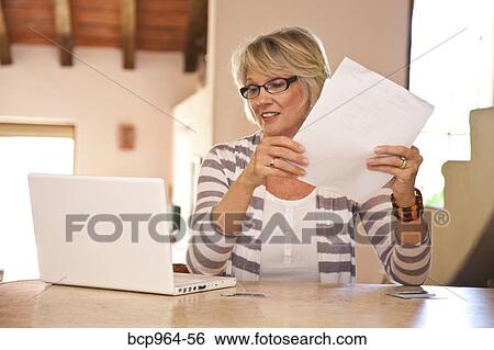 Stock Photograph - Woman at home office holding bills and looking at laptop. Fotosearch