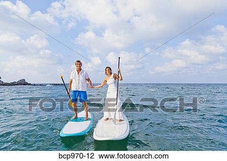 Stock Image - Dressed up man and woman riding paddle boards. Fotosearch