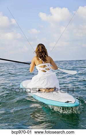 Stock Image - Woman in white dress riding paddle board. Fotosearch