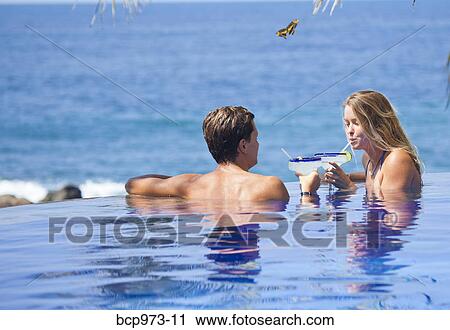 Stock Image - Young couple with cocktails in infinity pool. Fotosearch