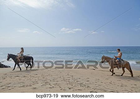 Stock Photo - Young man and woman riding horses on beach. Fotosearch