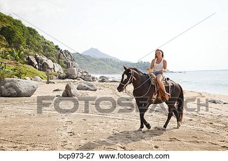 Stock Photo - Young woman riding horse on beach. Fotosearch