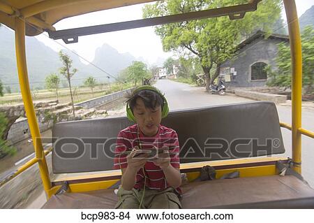 Picture - Boy wearing headphones uses an electronic device while riding a village jitney. Fotosearch