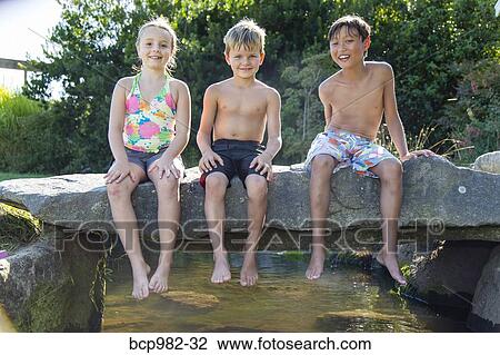 Stock Image - Three children sitting on a stone bridge over a small creek. Fotosearch