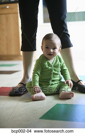 Picture - A baby sitting on kitchen floor with his mom behind him.. Fotosearch