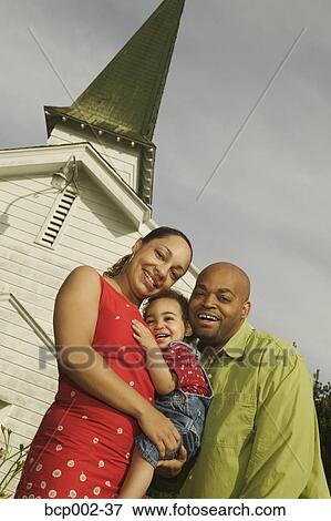Stock Photo - A family standing close together in front of white church. . Fotosearch