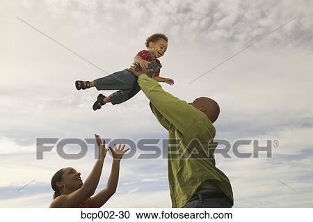 Stock Image - African-American father lifts his son into a cloudy white sky, with mother close by. . Fotosearch