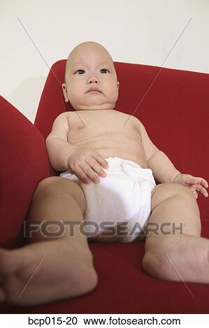 Stock Image - A baby seated on a red sofa and wearing a diaper. . Fotosearch
