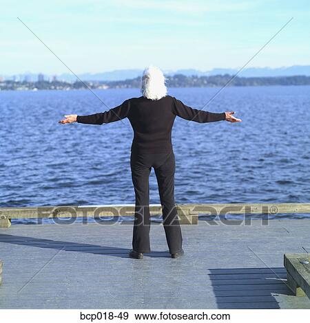 Stock Photo - A senior woman standing on a dock with her arms outspread.. Fotosearch