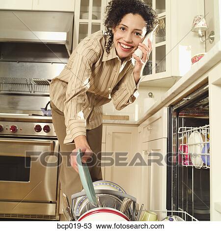 Stock Image - Young woman talking on the phone while loading her dishwasher.. Fotosearch