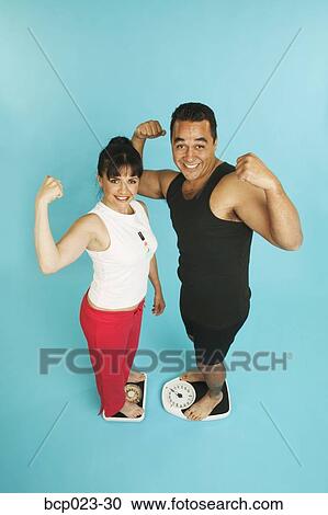 Stock Image - Couple standing on scales and flexing their muscles. Fotosearch