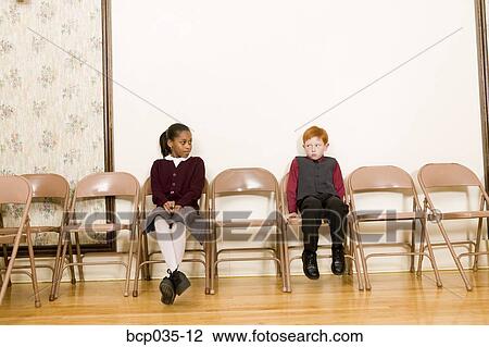 Stock Image - Girl and boy sitting in chairs against wall. Fotosearch