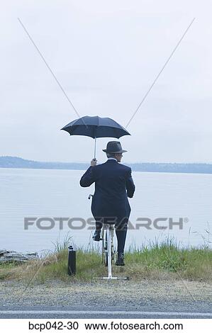 Stock Image - Businessman riding stationary bike near lake. Fotosearch