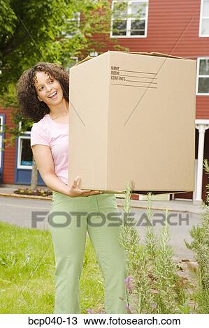 Stock Image - Smiling woman carrying large box. Fotosearch