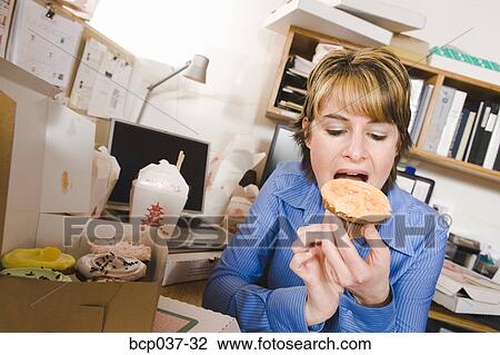 Stock Image - Office worker eating at her desk. Fotosearch