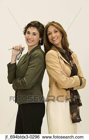 Stock Photograph - Two young businesswomen standing back to back looking at camera smiling. Fotosearch