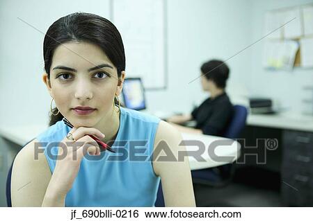 Stock Photograph - Young woman sitting in an office looking at camera with a young man sitting behind. Fotosearch