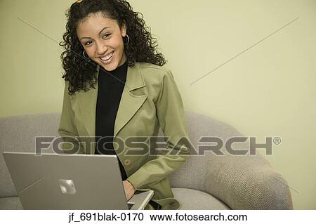 Stock Image - Young woman sitting on a couch working on a laptop smiling. Fotosearch
