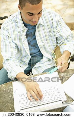 Stock Photo - High angle view of a young man sitting at a table in front of a laptop holding a credit card. Fotosearch