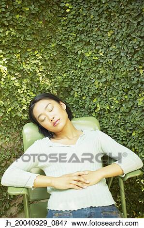 Stock Photo - High angle view of a young woman sitting on a chair outdoors with her eyes closed. Fotosearch
