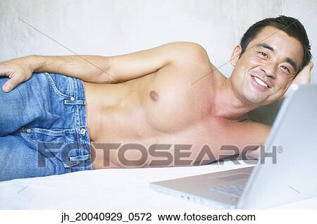 Stock Image - Mid adult man lying on a bed operating a laptop. Fotosearch