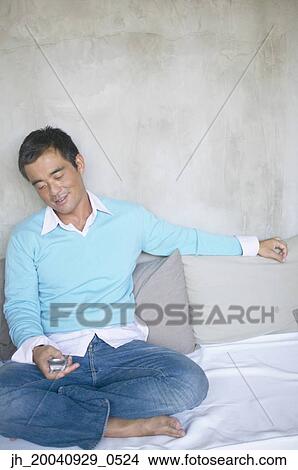 Mid adult man sitting on a bed operating a mobile phone View Large Photo Image Picture - Mid adult man sitting on a bed operating a mobile phone. Fotosearch