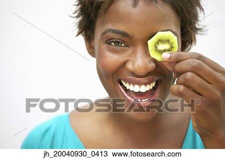 Portrait of a young woman holding a slice of kiwi fruit over her eye smiling View Large Photo Image Stock Image - Portrait of a young woman holding a slice of kiwi fruit over her eye smiling. Fotosearch