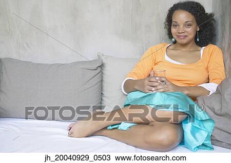 Portrait of a young woman sitting on a bed holding a glass View Large Photo Image Stock Image - Portrait of a young woman sitting on a bed holding a glass. Fotosearch