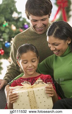 Stock Image - Young couple and their child holding a gift with a Christmas tree in the background. Fotosearch
