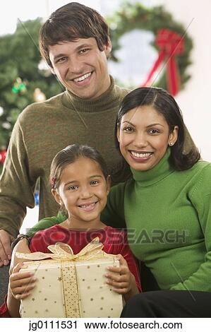Stock Image - Young couple and their child looking at camera smiling with a Christmas tree in the background. Fotosearch