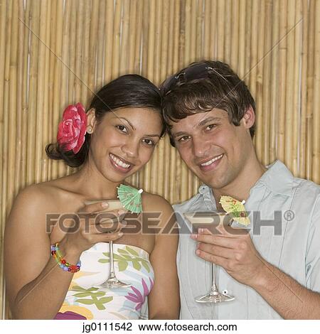 Stock Image - Young couple holding cocktails looking at camera. Fotosearch
