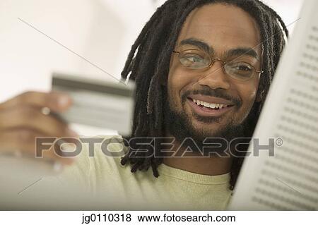 Stock Photo - Young man holding a credit card sitting in front of a computer screen. Fotosearch