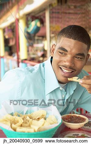 Young man sitting at a table eating View Large Photo Image Stock Photo - Young man sitting at a table eating. Fotosearch