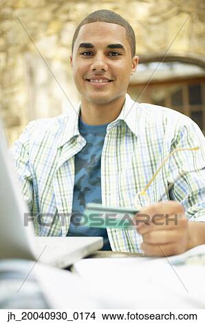 Young man sitting at a table in front of a laptop holding a credit card View Large Photo Image Picture - Young man sitting at a table in front of a laptop holding a credit card. Fotosearch