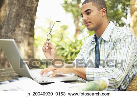 Young man sitting at a table working on a laptop View Large Photo Image Stock Image - Young man sitting at a table working on a laptop. Fotosearch