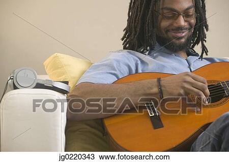 Young man sitting on a couch playing a guitar View Large Photo Image Stock Image - Young man sitting on a couch playing a guitar. Fotosearch
