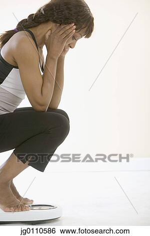 Stock Photograph - Young woman crouching on a weighing scale. Fotosearch