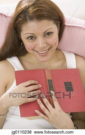 Young woman lying on a bed holding a book smiling View Large Photo Image Stock Photo - Young woman lying on a bed holding a book smiling. Fotosearch