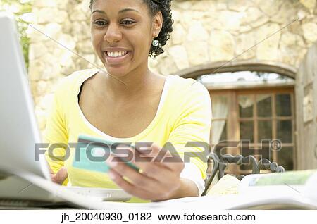 Young woman sitting at a table in front of a laptop holding a credit card smiling View Large Photo Image Stock Image - Young woman sitting at a table in front of a laptop holding a credit card smiling. Fotosearch